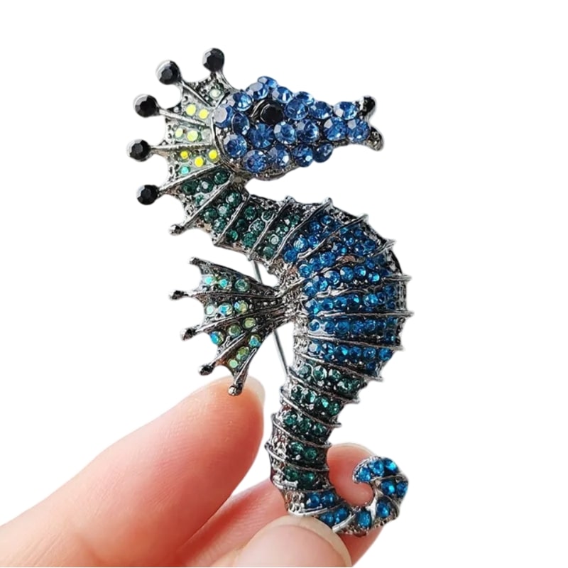 hand clutching a silver seahorse pin embellished with rhinestones in shades of lime, emerald, deep blue, and black